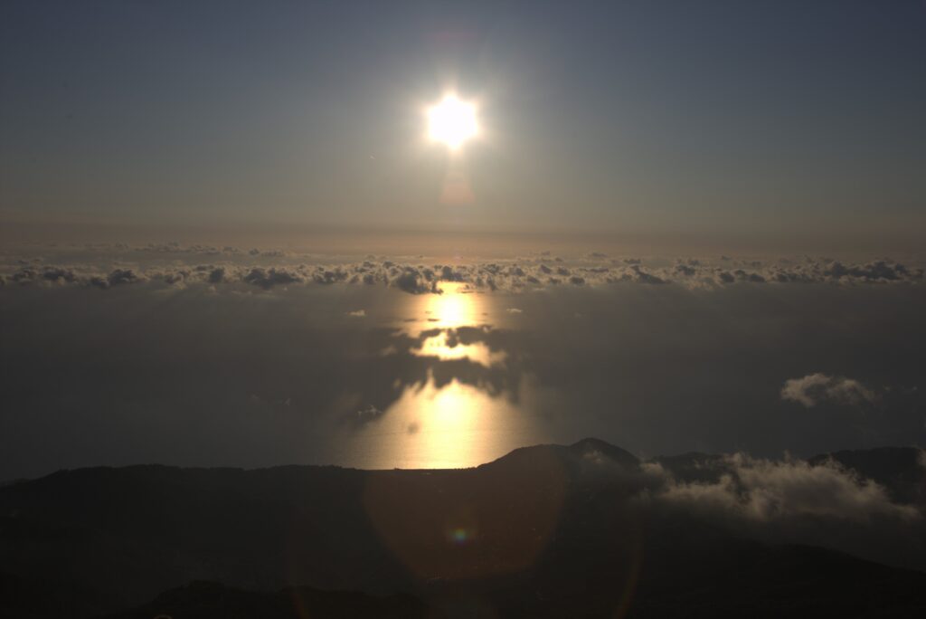 Inizio del tramonto: il sole ancora a metà del cielo, una striscia d'oro sul mare e le nuvole all'orizzonte.